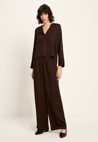 Frau mit kurzem, welligem Haar, die ein lockeres, dunkelbraunes V-Ausschnitt-Oberteil und weite Hose trägt, steht vor einem einfarbigen, hellen Hintergrund.