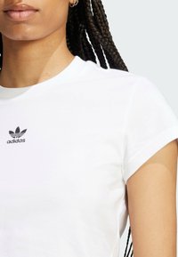 Biela tričko s okrúhlym výstrihom vyrobené z bavlny, s krátkymi rukávmi a čiernym logom Adidas trefoil na ľavej hrudi. Hladká textúra.