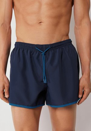 Torace maschile atletico che indossa pantaloncini da bagno blu navy con rifiniture turchesi e coulisse, in piedi davanti a uno sfondo semplice.