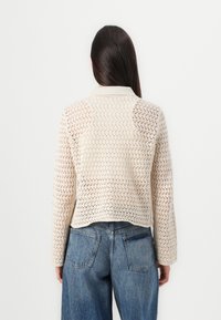 Gebreid cardigan in beige met een gestructureerd open steekpatroon. Kenmerken zijn een kraag en lange mouwen, gecombineerd met hoogwaisted denim jeans.