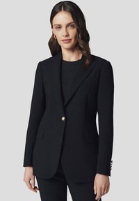 Czarna marynarka wykonana z teksturowanej tkaniny, z pojedynczym złotym guzikiem, klapami w szpic oraz przednimi kieszeniami. Stylizowana na dopasowaną sylwetkę.