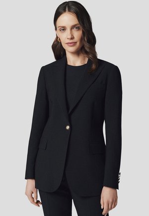 Czarna marynarka wykonana z teksturowanej tkaniny, z pojedynczym złotym guzikiem, klapami w szpic oraz przednimi kieszeniami. Stylizowana na dopasowaną sylwetkę.
