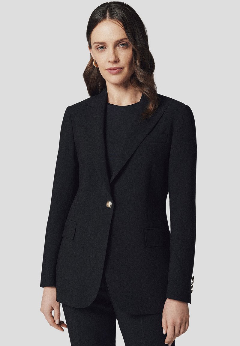 Czarna marynarka wykonana z teksturowanej tkaniny, z pojedynczym złotym guzikiem, klapami w szpic oraz przednimi kieszeniami. Stylizowana na dopasowaną sylwetkę.