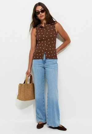 Kvinde iført brun ærmeløs prikket top, lyseblå vide jeans, mørke flade sko, solbriller og holder en flettet stråtaske.