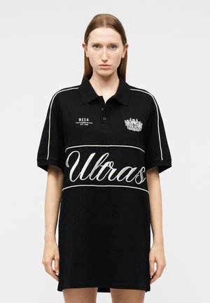 Γυναίκα που φοράει υπερμεγέθη μαύρο φόρεμα πόλο με λευκή επένδυση, κεντημένο κείμενο "Ultras" και έμβλημα στο στήθος, στέκεται μπροστά σε απλό φόντο.