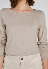 Beige långärmad topp med vid halsringning, tillverkad av mjukt tyg. Har ribbad struktur vid ärmsluten, tillsammans med ljusa byxor.