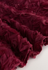 Bordeauxkleurige stof met gelaagde, driedimensionale bloemapplicaties gemaakt van een textuurrijke, lichtgewicht materiaal met rafelige randen.
