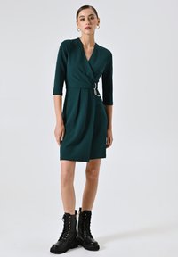Groene wikkeljurk met driekwartmouwen, zilveren ringdetail op de taille en een geplooide rok. Gepaard met zwarte veterschoenen tot de enkel.