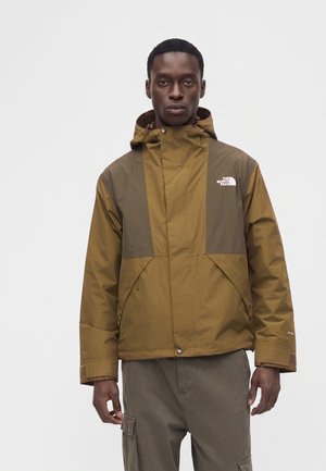 Homme portant une veste The North Face bicolore marron et vert olive avec capuche, debout sur un fond blanc uni.
