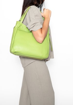 Vrouw in beige outfit draagt een grote limegroene leren handtas over haar schouder tegen een witte achtergrond.