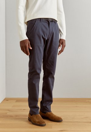 Pantalones chinos de algodón azul marino con corte slim, trabillas para cinturón, bolsillos frontales y costuras sutiles. Combinados con botas de ante marrón.