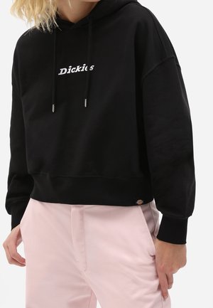 Persoon draagt een zwart kort hoodie met de tekst "Dickies" en lichtroze broek, staat met één hand dichtbij de zak.
