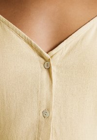 Gros plan sur une chemise beige à col en V avec deux petits boutons blancs sur un tissu texturé.