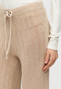 Μπεζ πλεκτό παντελόνι με κάθετες ρίγες και ελαστική μέση με κορδόνι, συνδυασμένο με λευκή ριγέ μπλούζα με μακριά μανίκια, χέρι ξεκουράζεται στον μηρό.