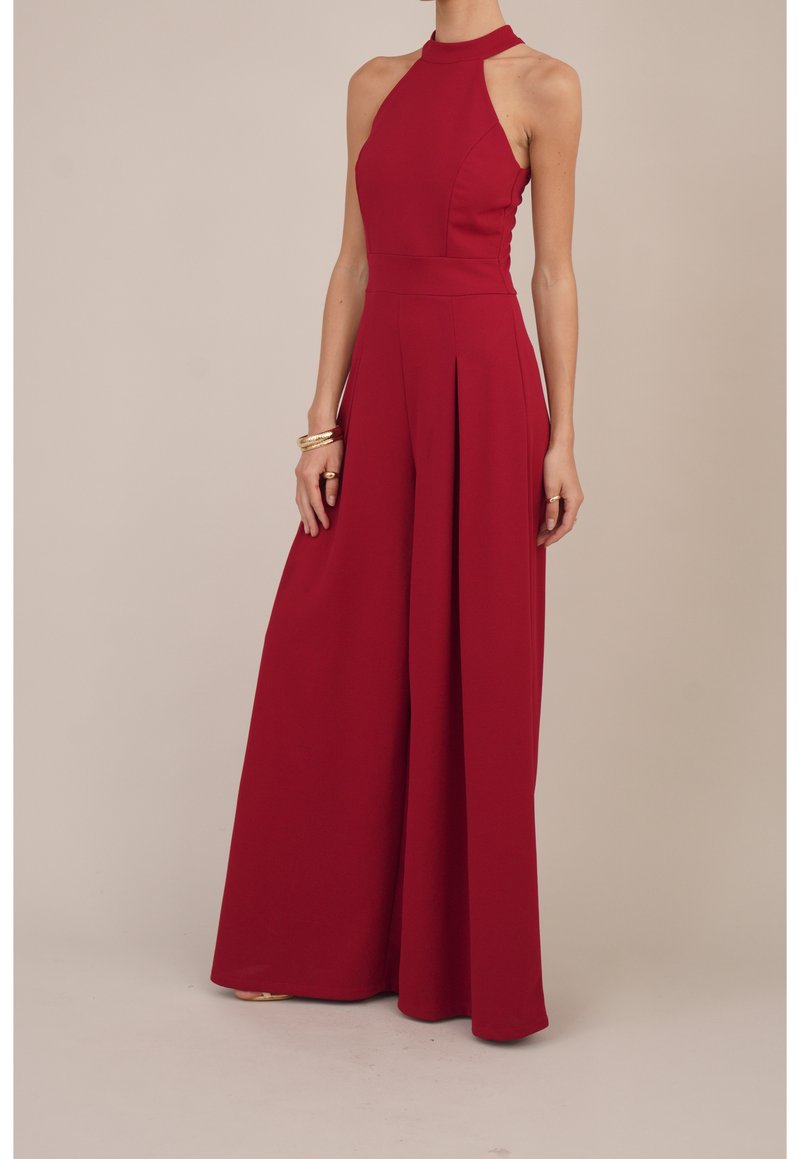 Combinaison rouge sans manches à col halter avec pantalon à jambes larges, portée avec des bracelets et des bagues dorés sur un fond neutre.