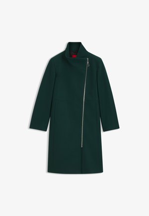 Manteau vert foncé à mi-cuisse avec des manches longues, fermeture éclair frontale asymétrique et col haut, posé à plat sur un fond blanc.