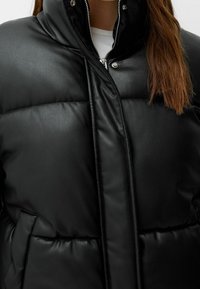 Närmast av en person som bär en svart vadderad imitationsläderjacka med dragkedja och tryckknappslås över en vit skjorta.