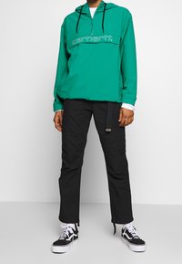 Teal grøn hættetrøje med halv lynlås og hvidt logo på brystet, kombineret med sorte cargobukser og sorte platformsneakers.