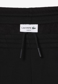 Svarta slim-fit sweatpants med dragsko i midjan, med en vit etikett märkt "LACOSTE" och en grön krokodil-logotype. Mjuk tygtextur.