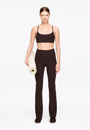 Czarny sportowy stanik i czarne rozszerzane legginsy z elastycznego materiału. Modelka trzyma zwiniętą matę do jogi na gładkim, neutralnym tle.