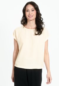 Gaiši bēša īsroku blūze ar apaļu apkakli un mīkstu, gludu tekstūru. Topiņš sniedzas līdz tieši virs gurniem un tam ir uzrollētas piedurknes.