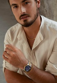 Eine elegante Uhr mit einem blauen Zifferblatt, einem Gehäuse aus Edelstahl in Silber und Gold und einem strukturierten goldenen Gliederarmband, das an einem unbedeckten Handgelenk getragen wird.