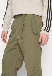 Lättviktsbyxor i olivgrönt med dragsko i midjan, utrustade med sidfickor och diskret logotypsbroderi. Slät textur och avslappnad passform.