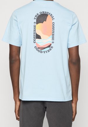 Jasnoniebieski bawełniany t-shirt z zaokrąglonym dekoltem, zdobiony kolorową grafiką na plecach z tekstem, falami i abstrakcyjnymi kształtami.