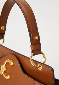 Brown leather handbag with a curved handle, golden hardware, and contrasting stitching. Features a distinctive logo and round metal ring accents.