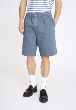 Mand iført blå shorts i mellemlængde med elastisk talje og knap, kombineret med hvide sokker og sorte loafers, stående mod en ensfarvet baggrund.