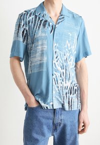 Koszula na krótki rękaw na guziki w jasnoniebieskim kolorze, z abstrakcyjnymi białymi i czarnymi wzorami oraz luźnym krojem. Wykonana z lekkiego materiału.