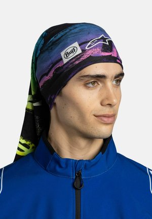 Giovane uomo che indossa una fascia colorata con il logo Buff e una giacca blu con zip, guarda leggermente verso destra su uno sfondo semplice.