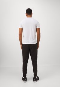 Witte sportieve T-shirt met korte mouwen, gecombineerd met zwarte, taps toelopende broek en zwarte schoenen, gezien vanaf de achterkant tegen een lichte achtergrond.