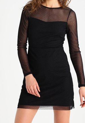 Femme portant une robe noire ajustée avec des manches longues en tulle transparent à pois, détails sur le haut de la poitrine et l'ourlet.