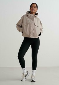 Donna con giacca beige con cappuccio, leggings neri, calzini bianchi e scarpe da corsa grigie, in piedi contro un muro bianco semplice.