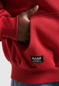 Červená mikina s kapucňou, s rebrovanými manžetami a pásom. Obsahuje predné vrecko a čierny štítok so slovami "KANI THE ORIGINAL EST. 1989."