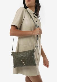 Borsa a tracolla verde in pelle con texture circolare in rilievo, hardware argentato e un accent logo. È indossata con un vestito beige a bottoni.