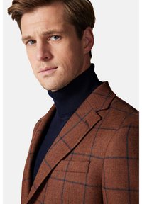 Brązowa marynarka w kratę o teksturowym wykończeniu, zestawiona z dopasowanym granatowym golfem. Posiada sztywne klapy i szczegóły kieszeni.