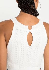 Top cropped branco em malha com um padrão de zigue-zague, decote redondo e detalhe nas costas com abertura em forma de chave, segurado por um botão.