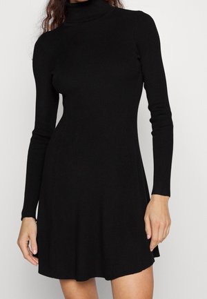 Vrouw draagt een zwarte ribgebreide jurk met lange mouwen, een hoge col en een uitlopende zoom, getoond van middel tot nek.