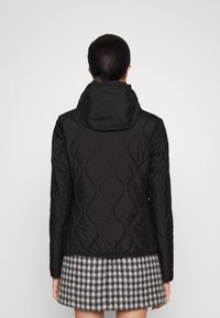 Sort quiltet jakke med hætte, der har et struktureret mønster. Blødt stof og tætsiddende design, båret over en grå og sort ternet nederdel.