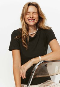 T-shirt noir à manches courtes, en coton lisse. Le mannequin porte un collier en coquillage et plusieurs bracelets, assis sur une chaise en chrome.