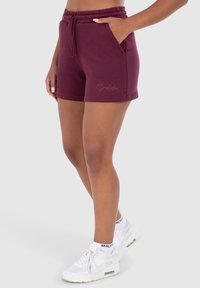 Bermuda maroon dengan pinggang elastis dan tali serut, dilengkapi dengan saku samping dan logo teks kecil di bagian hem. Dipadukan dengan sepatu sneaker putih.