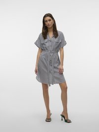 Robe chemise rayée en bleu marine et blanc avec des manches courtes, une patte de boutonnage, deux poches poitrine et une taille nouée. Associée à des sandales à talons vertes.