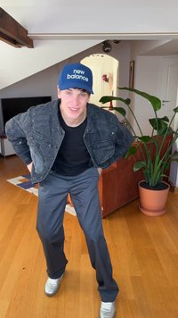 Casaco de ganga preto com design texturizado, camiseta preta, calças cinzentas e ténis prateados. Boné azul com texto "new balance". Ambiente interior.