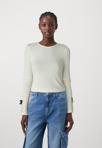 Maglietta a maniche lunghe verde chiaro realizzata in tessuto a coste con scollo rotondo e dettagli neri ai polsini, abbinata a jeans blu larghi.