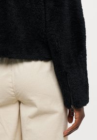 Schwarz plüschige Jacke mit strukturierter Oberfläche, mit einem Reißverschluss am Ärmel und über hellbeigen Cordhosen getragen.