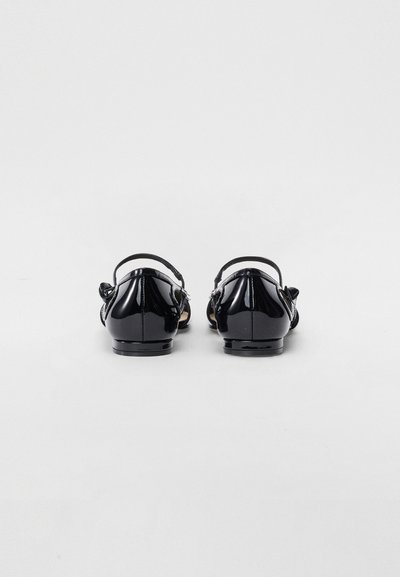 Chaussures en cuir verni noir à petit talon, dotées d'une sangle de cheville et d'un accent décoratif en forme de nœud à l'arrière. Texture lisse et brillante.