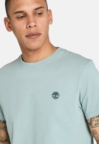 Ljusblå bomulls t-shirt med rund halsringning och korta ärmar; har en mörkgrön broderad trädlogotyp på bröstet.