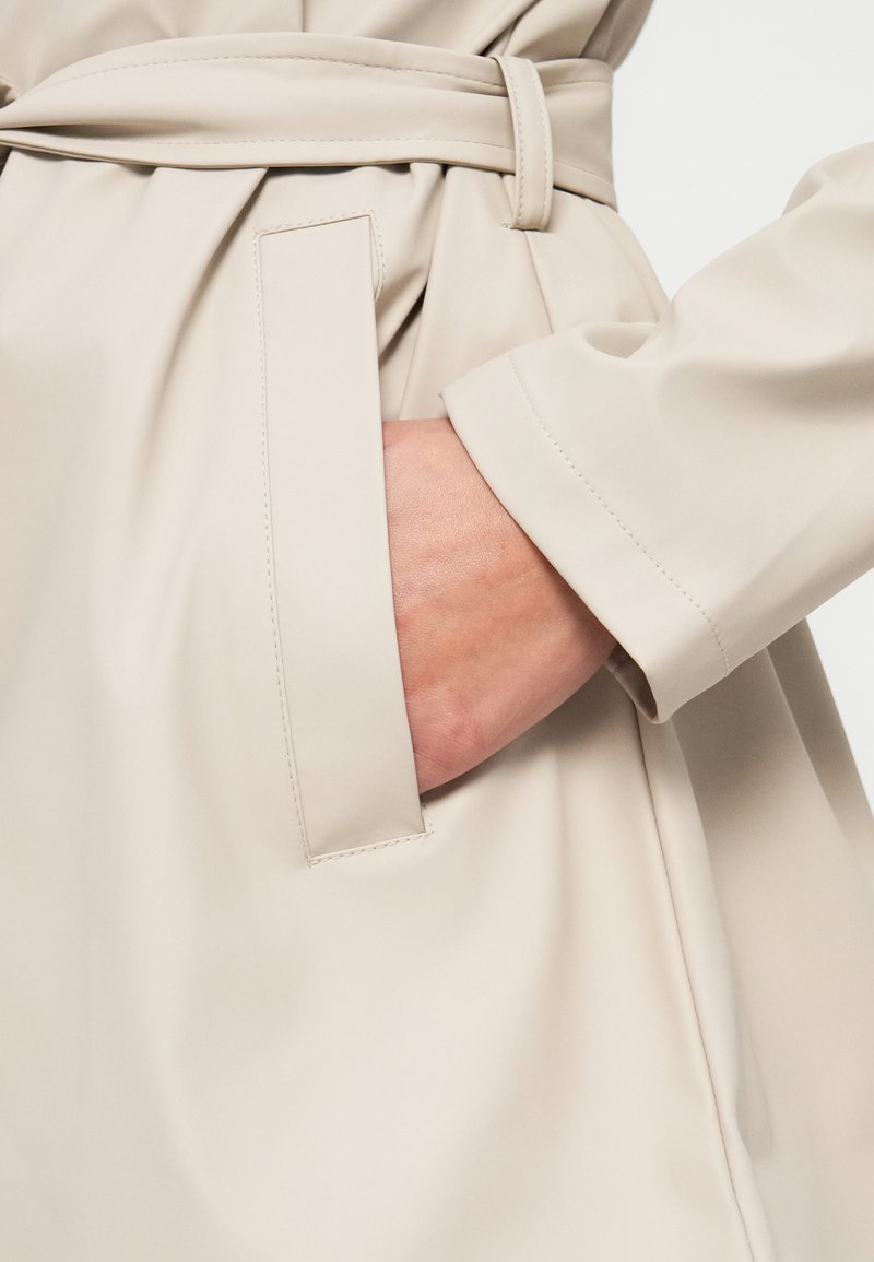 Abrigo trench beige con cinturón en la cintura y un bolsillo lateral, que presenta un material liso y brillante y costuras precisas, destacando un diseño minimalista.